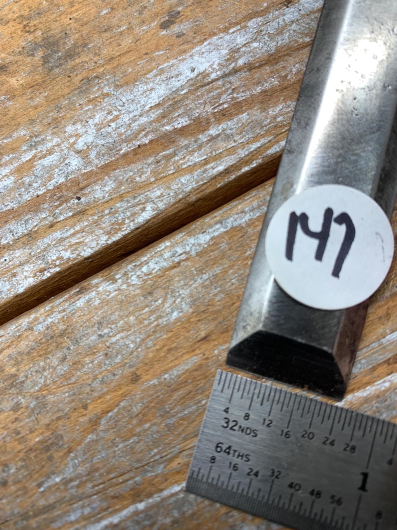 May include: A metal chisel with a beveled edge, next to a metal ruler. The chisel has a sticker with the number 147 on it. The ruler shows measurements in inches and fractions.