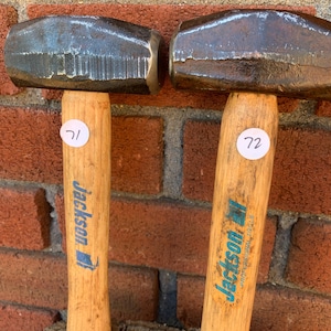 May include: Two sledgehammers with wooden handles and dark metal heads. The handles have the blue text "Jackson Professional Tools". Each handle has a white sticker with a number. The background is a red brick wall.