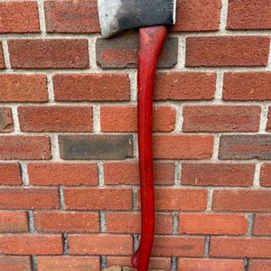 May include: A felling axe featuring a dark, weathered metal head with a sharp, worn edge and a long, smooth, vibrant red wooden handle. The axe is positioned vertically, with the base of the handle resting on a circular, textured wooden log.