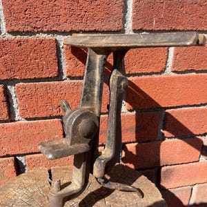 May include: A vintage cast iron cobbler shoe stand featuring a dark, weathered metal finish with a horizontal platform, a vertical support arm, and a threaded screw clamp mechanism at the base for securing the tool to a workbench surface.