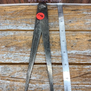 May include: A vintage metal outside caliper tool with a dark, weathered patina and a central pivot bolt. A bright orange circular sticker with the number 100 is attached to the upper arm. A metal ruler is placed alongside, showing a length of 8 inches.