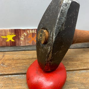 May include: A heavy-duty, vintage-style hammer with a wooden handle and a round, wooden head. The hammer is resting on a red, rubber mallet.