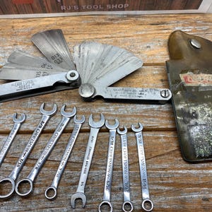May include: A collection of vintage tools, including open-end wrenches and a feeler gauge set, arranged on a wooden surface. The wrenches are silver-toned, and the feeler gauge is made of thin metal blades. A leather case is also present.