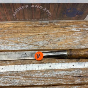 May include: A metal chisel with a wooden handle, resting on a wooden surface. A metal ruler is placed below the chisel. The chisel has a blade length of approximately 11 inches.