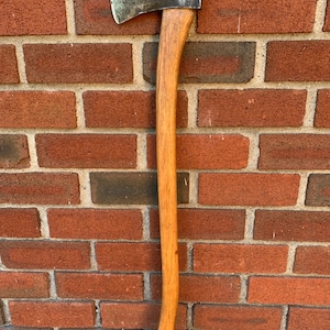 May include: A felling axe with a curved wooden handle and a metallic head. The axe head features a dark, weathered steel finish with a sharp, slightly curved cutting edge. The handle is made of light brown wood with a visible grain pattern.