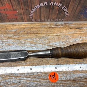 May include: A wood chisel with a dark wooden handle and a metal blade. The chisel is placed on a wooden surface next to a ruler. The text "Hammer and Edge" and "RJ's Tool Shop" is visible in the background.
