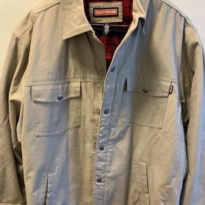 May include: A tan work jacket with a red and black plaid lining. The jacket has a button closure, two chest pockets, and two lower pockets. The label on the jacket reads "CRAFTSMAN".