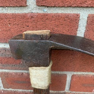 May include: A vintage pickaxe featuring a dark, weathered metal head with a visible wooden wedge at the top. The handle is made of wood and is reinforced with a thick, light-colored twine wrapping just below the metal head of the tool.