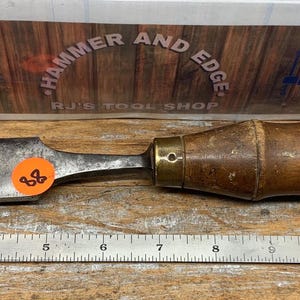 May include: A vintage wood-handled chisel with a metal blade. The chisel has a worn, aged appearance, with a wooden handle and a metal blade. The blade is approximately 10 inches long. The background features a sign that reads "Hammer and Edge".