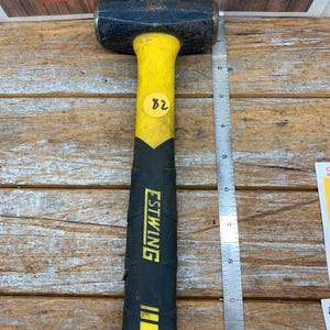 May include: A blue metal sledgehammer with a yellow and black rubberized handle featuring the Estwing brand name and a yellow sticker with the number 82. A metal ruler placed alongside shows the tool measures approximately 10.5 inches in length.