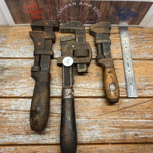 May include: Three vintage metal pipe wrenches with dark wooden handles arranged vertically. The center wrench features a small white circular tag with the number 18. A metal ruler is positioned to the right, showing a length of 5 inches.