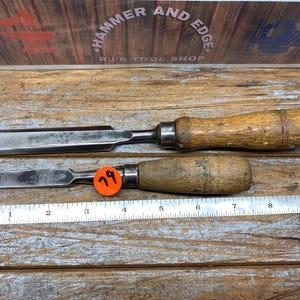 May include: Two vintage wood-handled chisels with metal blades. One chisel has an orange price tag with the number 79. A metal ruler is placed below the chisels. The background features a wooden surface and a sign that reads "Hammer and Edge".