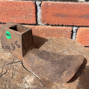 May include: A rusted iron adze head with a rectangular socket for a handle. The metal surface shows heavy oxidation and wear. A green circular sticker with the number 100 is attached to the side, along with the handwritten black text 3.5 lb.