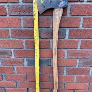 May include: A vintage axe with a wooden handle and a metal head. The axe head has a worn appearance with visible markings. A yellow ruler is placed next to the axe, showing a measurement of approximately 36 inches.