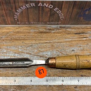 May include: A vintage wood chisel with a metal blade. The chisel has a light-colored wooden handle and a metal blade. The blade is silver and the handle is light brown. The chisel is lying on a wooden surface. The image also includes a ruler.