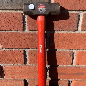 May include: A heavy-duty sledgehammer with a dark metal head and a bright red wooden handle. The handle is labeled "Plumb" in white lettering. The head has a sticker that reads "Plumb 48". The hammer is set against a red brick wall.