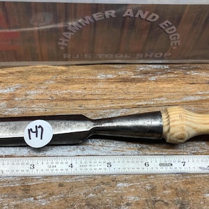 May include: A metal chisel with a wooden handle, resting on a wooden surface next to a ruler. The chisel has a dark metal blade and a light-colored wooden handle. A sticker with the number "17" is on the chisel.