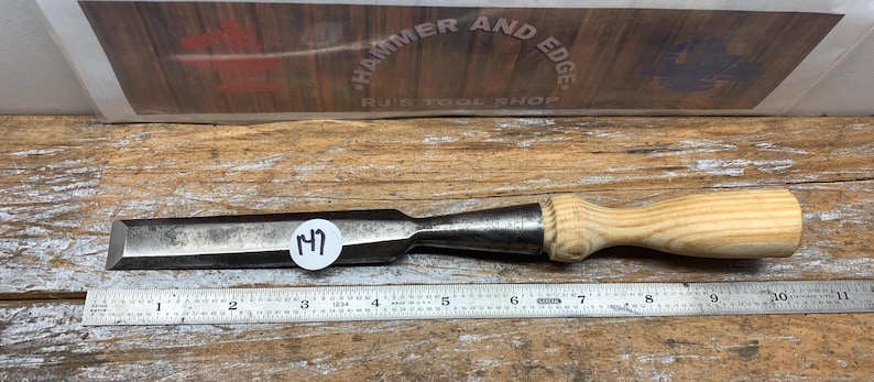 May include: A metal chisel with a wooden handle, resting on a wooden surface next to a ruler. The chisel has a dark metal blade and a light-colored wooden handle. A sticker with the number "17" is on the chisel.