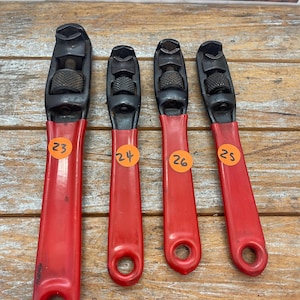May include: Four adjustable wrenches with black metal heads and red handles. Each tool features a knurled adjustment wheel and a circular hole at the base of the handle. Orange circular stickers with the numbers 23, 24, 26, and 25 are attached.