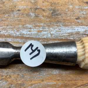 May include: A metal chisel with a wooden handle. The chisel has a silver blade and a light-colored wooden handle. A white sticker with the number "147" is affixed to the chisel.