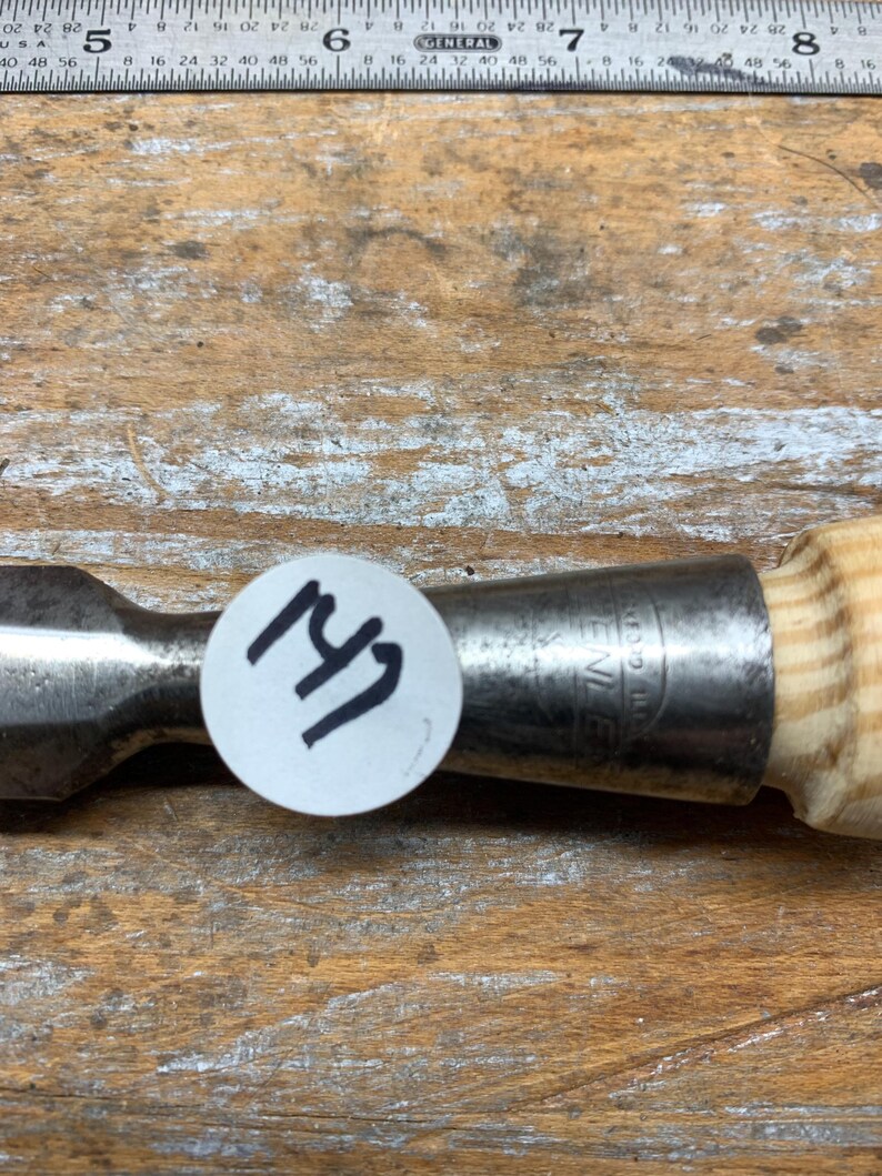 May include: A metal chisel with a wooden handle. The chisel has a silver blade and a light-colored wooden handle. A white sticker with the number "147" is affixed to the chisel.