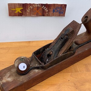 May include: A vintage wooden hand plane with a dark metal blade and handle. The plane is resting on a wooden surface. Above the plane is a sign that reads "R's Tool Shop" with images of tools.