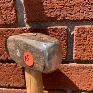 May include: A heavy-duty metal sledgehammer head with a weathered, dark grey finish and a polished, rounded striking face. A small, circular orange sticker with the handwritten number 85 is affixed to the side of the metal head, which is attached to a wooden handle.