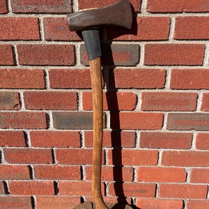 May include: A felling axe with a dark metal head and a long, curved wooden handle. The axe features a black rubberized grip section just below the metal head, which is securely attached to the wooden shaft. The metal head has a wide, sharp blade edge.