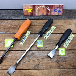 May include: Three metal pry bars with black and orange handles are displayed on a wooden surface. The tools are labeled with letters and numbers. A sign above reads "HIS TOOL SHOP".