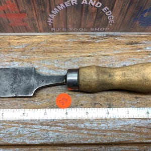 May include: A vintage woodworking chisel with a wooden handle and a wide, beveled blade. The blade shows signs of wear and age. A ruler is placed below the chisel, and the background features a sign that reads "Hammer and Edge, RJ's Tool Shop."