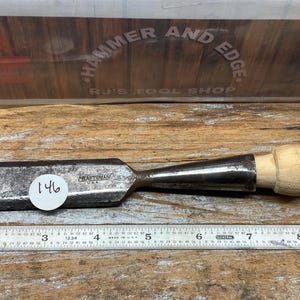 May include: A well-used wood chisel with a light-colored wooden handle and a metal blade. The blade is marked with the number "146" on a white sticker. A metal ruler is placed next to the chisel. The background features the text "HAMMER AND EDGE" and "R.J.'S TOOL SHOP".