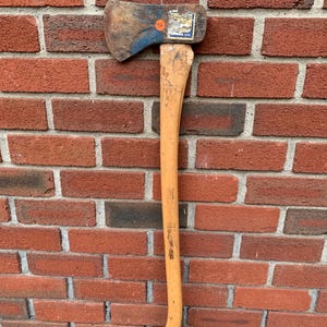 May include: A vintage felling axe with a weathered metal head featuring blue paint remnants and a worn wooden handle. A small orange circular sticker and a rectangular label with the text "PLUMB" and "CAMPING AXE" are visible on the metal head.