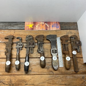 May include: Eight vintage adjustable wrenches with wooden handles, numbered 1 through 7, on a wooden surface. The wrenches are rusty and have a worn appearance.  The wrenches are arranged in a row from left to right.