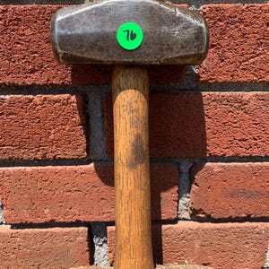 May include: A heavy-duty sledgehammer with a dark metal head and a wooden handle. A green sticker with the number 76 is affixed to the hammer head. The hammer is set against a red brick wall.