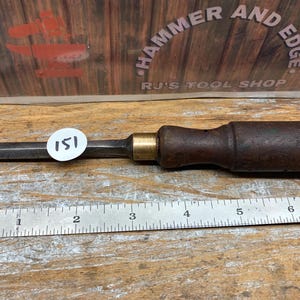 May include: A vintage wood chisel with a dark brown handle and a metal blade. The chisel has a brass ferrule and a round white sticker with the number 151 on it. A metal ruler is in the foreground.