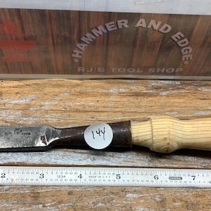 May include: A vintage wood chisel with a metal blade and a light-colored wooden handle. The blade shows signs of wear. A metal ruler is below the chisel. The background features a sign that reads "Hammer and Edge" and "RJ's Tool Shop."