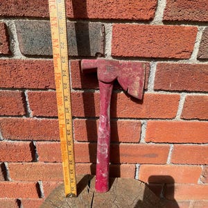 May include: A red-painted hatchet with a worn finish positioned next to a yellow wooden ruler. The ruler shows the hatchet head is aligned near the 23-inch mark and the handle extends down to approximately the 35-inch mark on the scale.
