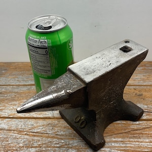 May include: A small, antique anvil with a pointed horn and a flat top, next to a green soda can. The anvil is made of dark metal and sits on a wooden surface. The can has nutrition facts printed on it.