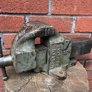 May include: A vintage Morgan Chicago bench vise with a weathered sage green finish and a metal handle. The cast iron tool features a sliding jaw mechanism and a prominent screw handle, showing signs of heavy use and surface wear on the metal body.