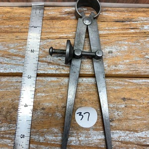 May include: A vintage metal caliper tool with a screw adjustment, used for measuring. A metal ruler is next to the caliper. The number "37" is written on a white sticker. The background is a wooden surface.