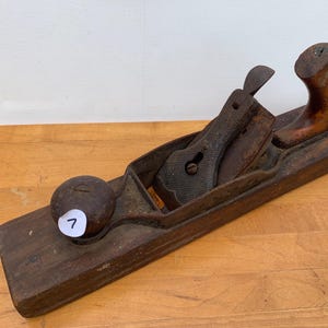 May include: A vintage wooden hand plane with a dark brown finish. The plane features a metal blade and a wooden handle. A white sticker with the number 7 is on the front. The tool is designed for woodworking and carpentry.