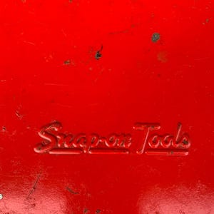 May include: A close-up of a bright red metal surface with the embossed text "Snap-on Tools". The surface has minor wear and tear, with small blemishes. The image is well-lit, highlighting the details of the text.