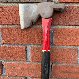 May include: A vintage Plumb hatchet with a red handle and a black Fiber-Glass handle. The axe head is silver and shows signs of wear. The hatchet is resting against a red brick wall.