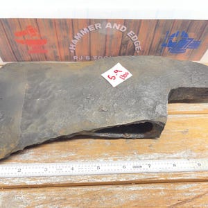 May include: A dark gray metal axe head with a textured surface and a central eye for a handle. A white sticker on the axe head displays the text 5.9 lbs. A metal ruler is positioned along the bottom edge, showing a length of approximately 10 inches.