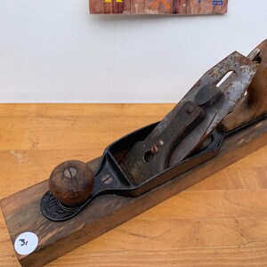 May include: A vintage wooden hand plane with a dark metal blade and a wooden handle. The plane has a rectangular wooden base and a round wooden knob. The blade is slightly rusted, indicating age and use. The tool is designed for woodworking.