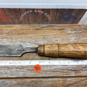 May include: A vintage wood-handled chisel with a metal blade. The chisel is placed on a wooden surface next to a ruler. The handle is a light brown color, and the blade is a darker, aged metal. The text "Hammer and Edge" is visible in the background.