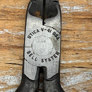 May include: Close-up of a pair of Utica V-41 USA wire cutters. The metal tool has a black finish with the text "UTICA V-41 USA BELL SYSTEM" printed on the silver-colored center. The handles are red.