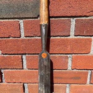 May include: A metal wood chisel featuring a long, flat, rectangular blade with a beveled edge and a cylindrical wooden handle capped with a metal ring. The tool has a small circular hole near the neck and an orange sticker with the number 141.