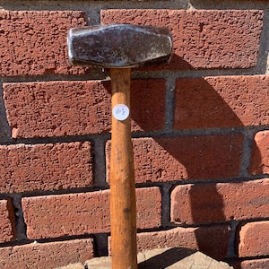 May include: A metal hammer with a wooden handle. The hammerhead is a dark, metallic colour, and the handle is a light brown. The hammer is resting on a wooden surface in front of a red brick wall. A small white sticker with the number 68 is on the handle.