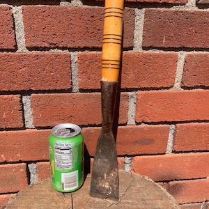 May include: A wooden-handled chisel with a metal blade, resting on a tree stump. The chisel is approximately 12 inches long and has a brown wooden handle with a light brown stripe pattern. The metal blade is rusty and has a slight curve.